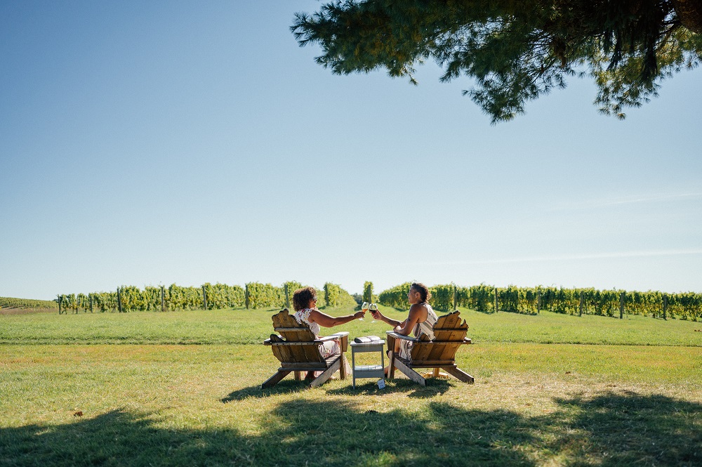two women sitting in wooden chairs overlooking the vineyard, cheering their wine glasses, sky is bright blue