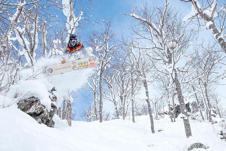 A snowboarder is in mid-air after riding off a rock formation on a snowy slope with blue skies above.