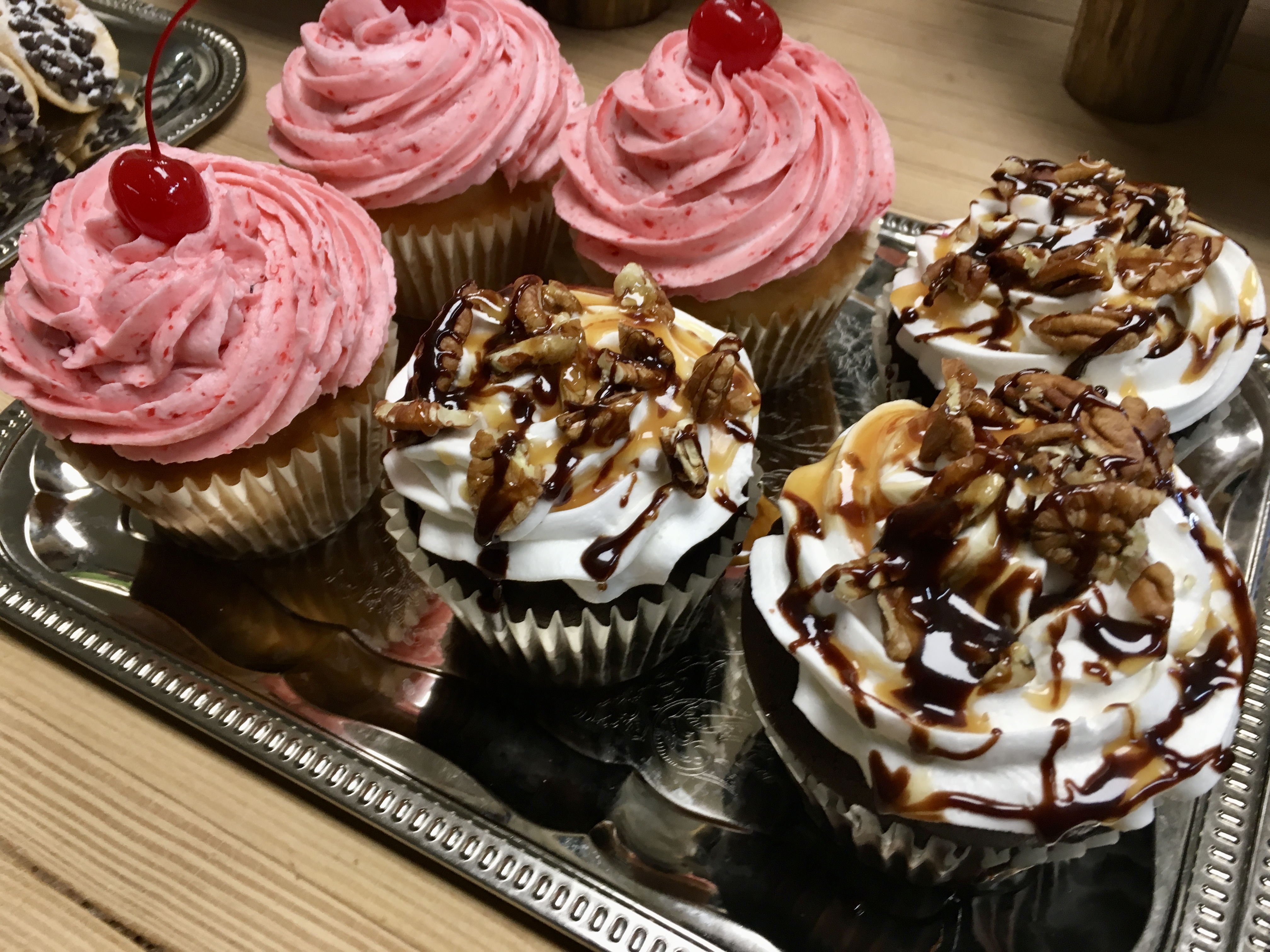six decorated cupcakes on a pan, 3 red with cherries on top and 3 chocolate with caramel and chocolate drizzled on top