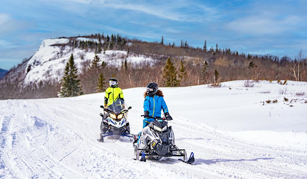 Two people riding snowmobiles on a snowy trail with a snow-covered mountain in the background.