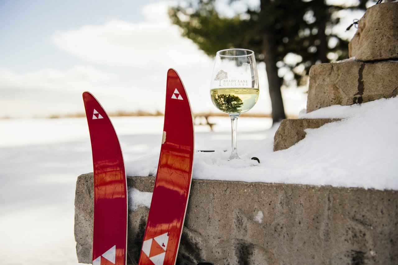 Shady Lane Cellars A pair of cross country skis leaning against a stone wall beside a glass of white wine.