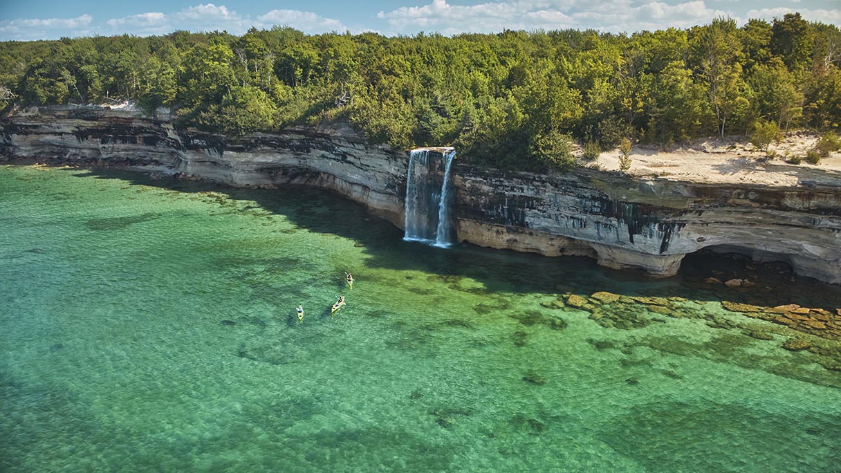 Kayakers on Lake Superior with a waterfall cascading off a tree-covered cliff.
