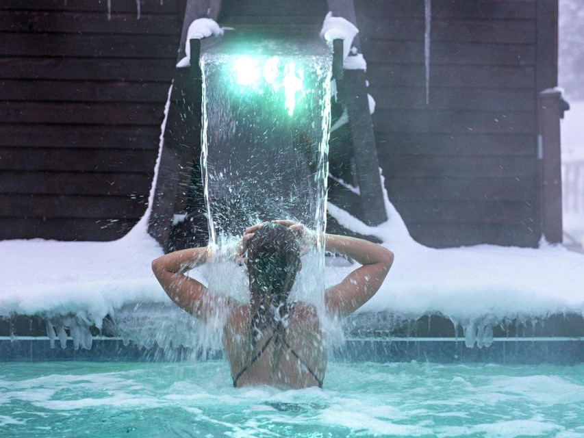 A woman under a water feature in an outdoor pool during winter.