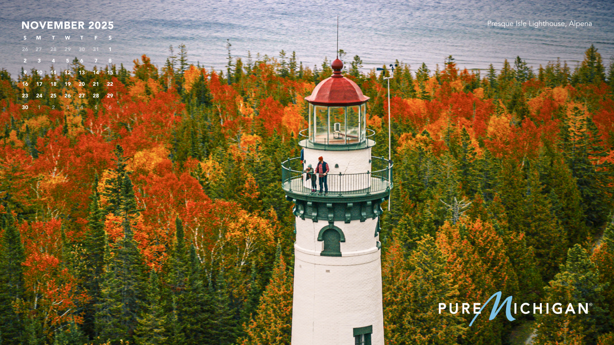 2025 Pure Michigan Calendar - November A November 2025 calendar graphic on a photo of a lighthouse above fall trees.