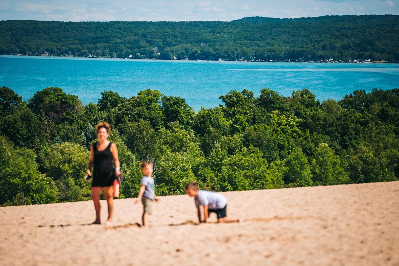 A woman and two children climbing a sand dune that overlooks an inland lake. 