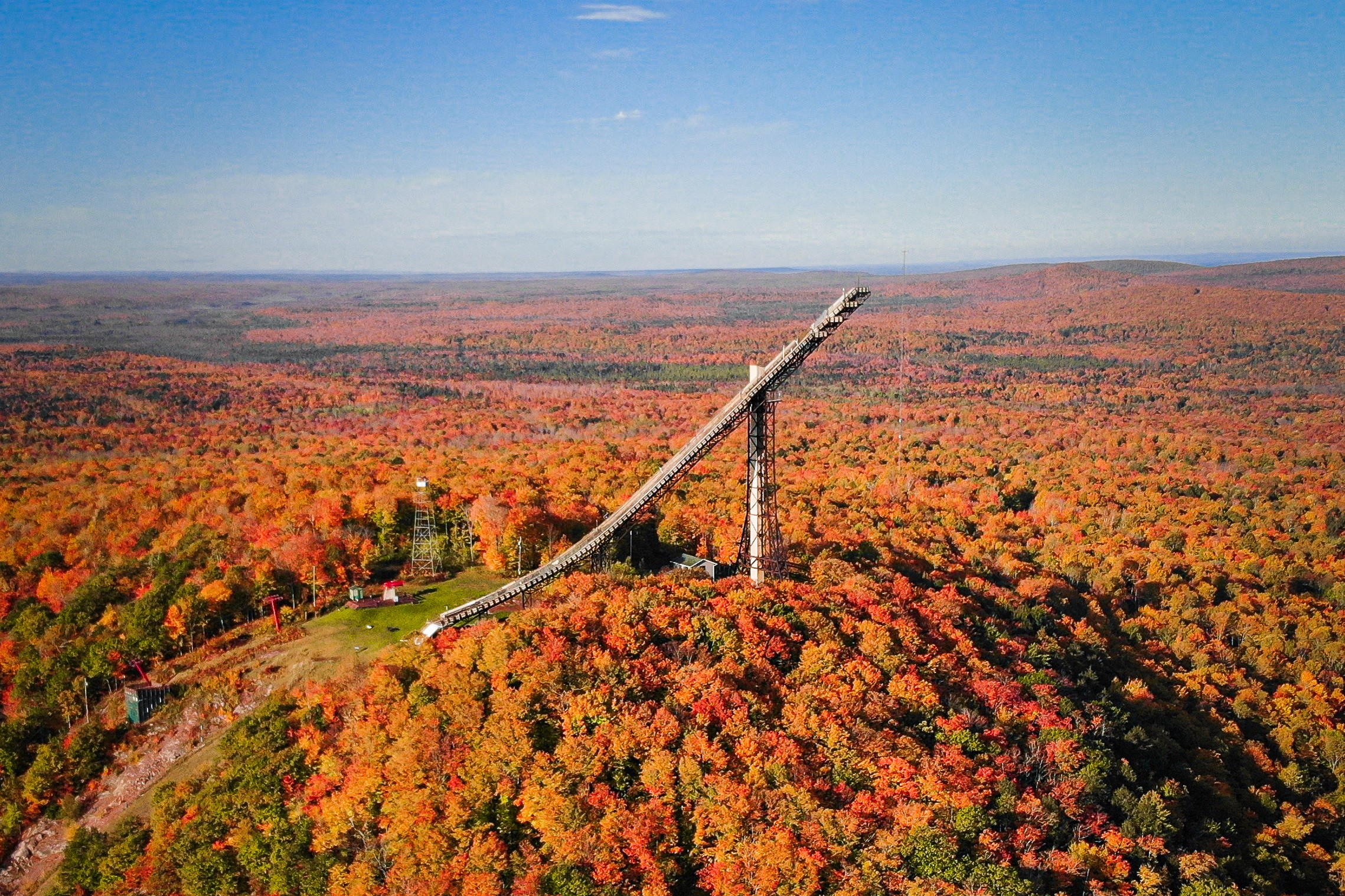 An artificial ski jump on a hill surrounded by tress displaying fall foliage. 