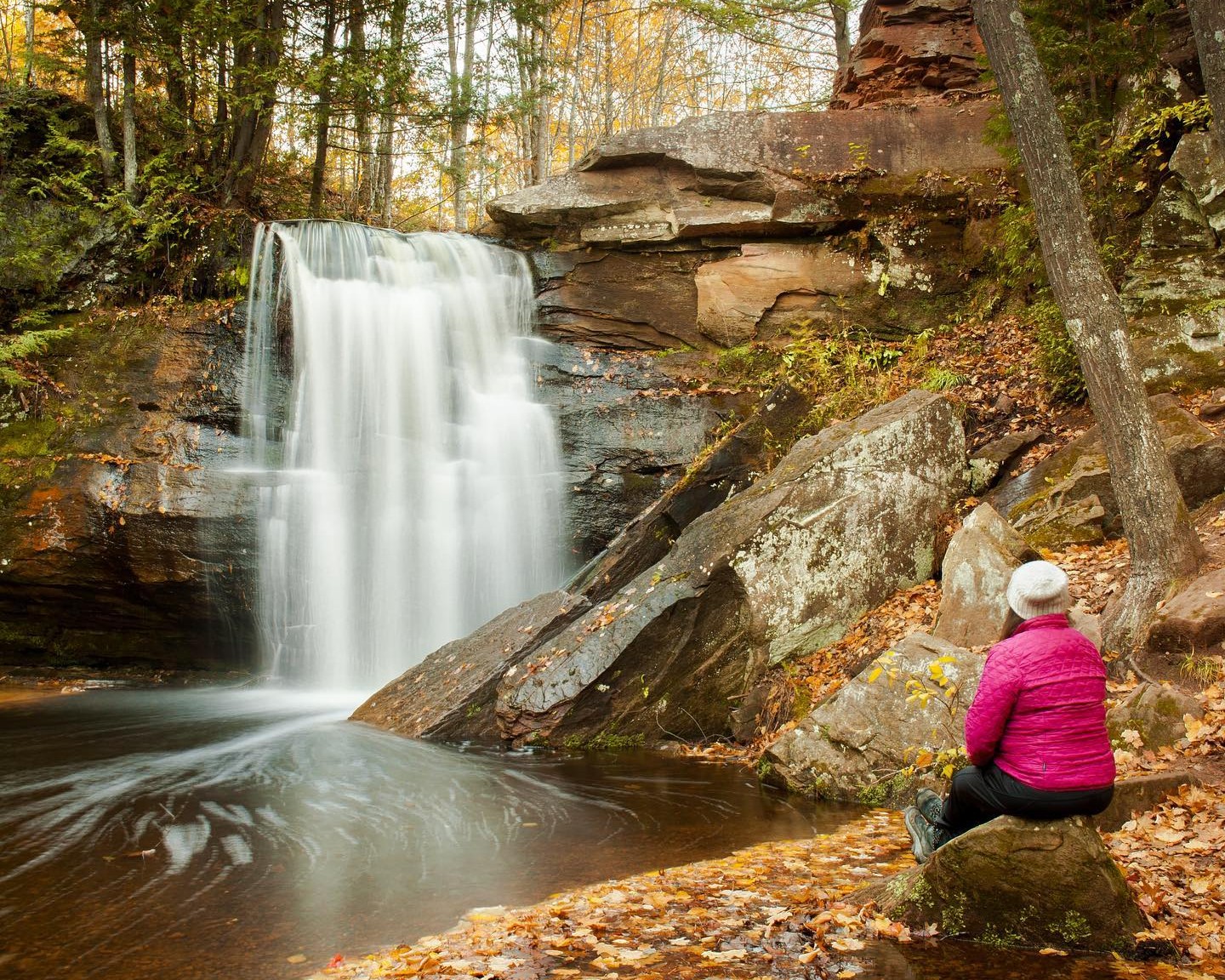 A woman sitting on a rock beside a waterfall during fall. 