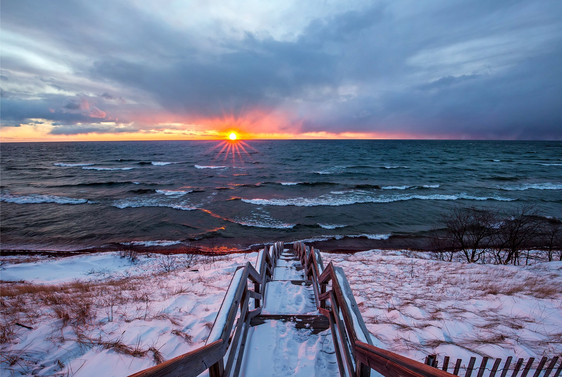 A wooden staircase leading down a snowy bluff to a wavy Lake Michigan at sunset. 