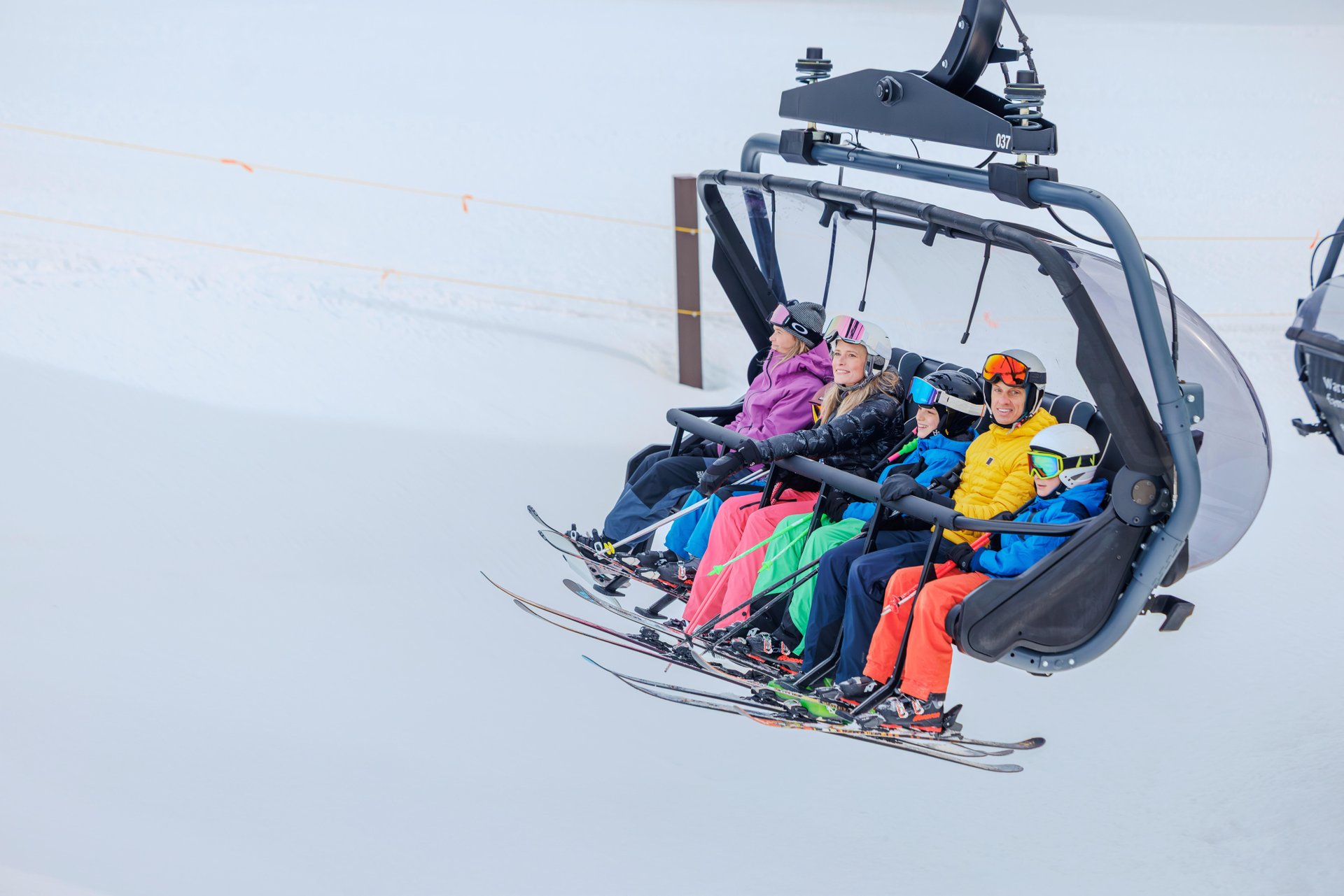 A family of five people on a partially enclosed chairlift, traveling up a mountain.