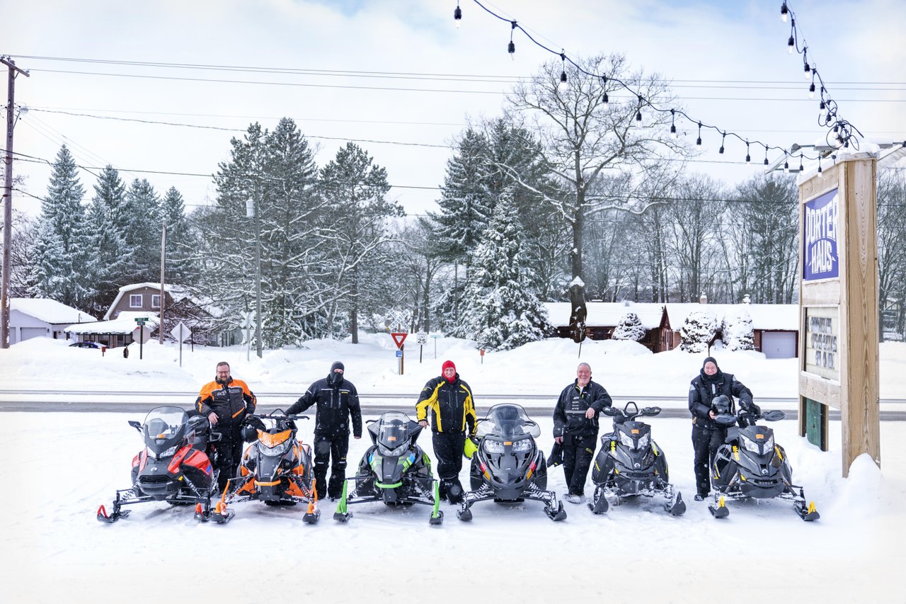 Five snowmobilers in winter gear standing beside their snowmobiles. 