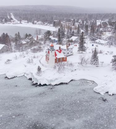 A lighthouse on a snow-covered and forested coastline overlooking an icy Lake Superior. 