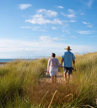 An elderly couple walking hand-in-hand along a grassy beach. 